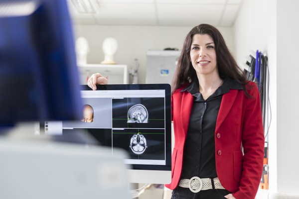 Silvia Daun leans with one arm over a screen and smiles friendly into the camera. The screen shows MRI scans of a human brain and a reconstruction of a head.